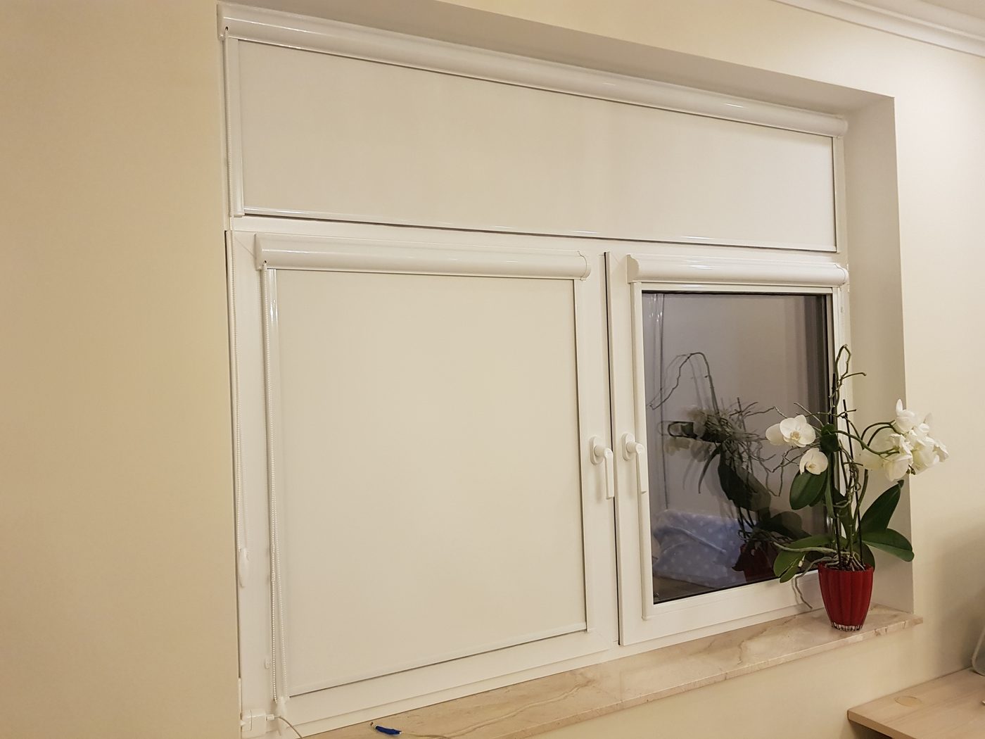 Baltas kasešu žalūzijas logā ar augšējo vērtni — kasete uz katras vērtnes